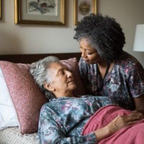 A compassionate caregiver gently leans toward an elderly woman resting in bed, providing comfort and support as part of hospice care Tulsa, OK in a warm home environment.